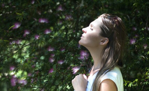 woman closing her eyes against sun light standing near purple petaled flower plant